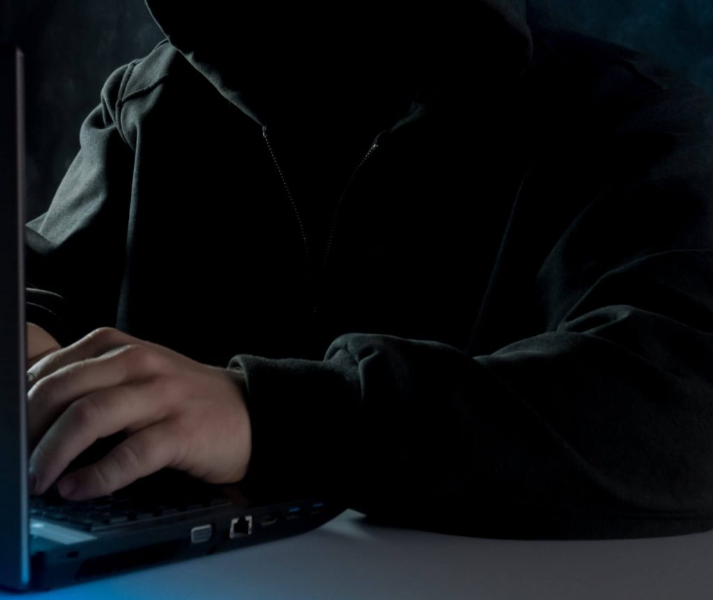 An anonymous person in dark clothing, sitting in front of a laptop with a mysterious aura.